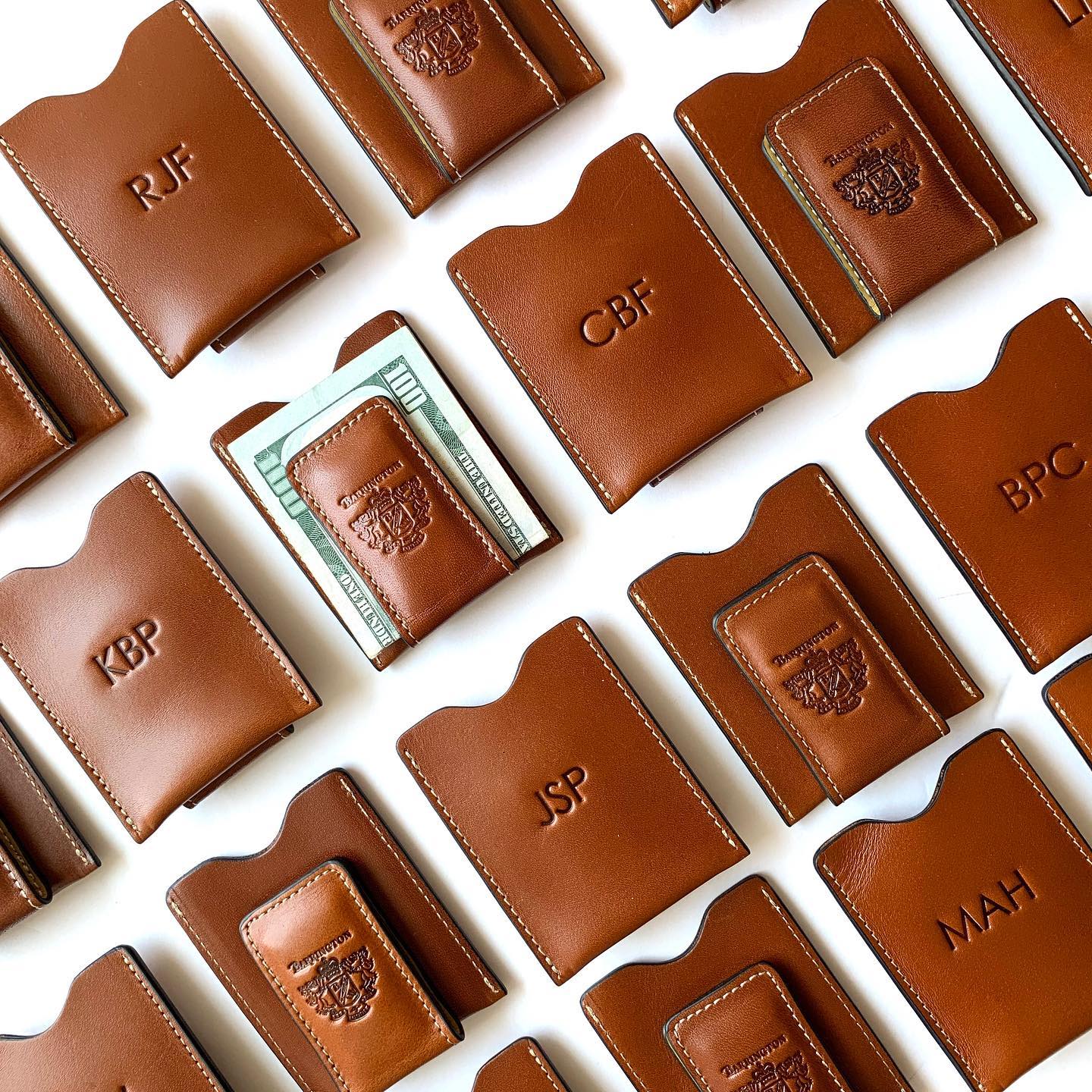 Multiple British tan leather money clips fanned out, each displaying unique custom monograms, highlighting personalization options for gifts and making each pocket wallet unique.