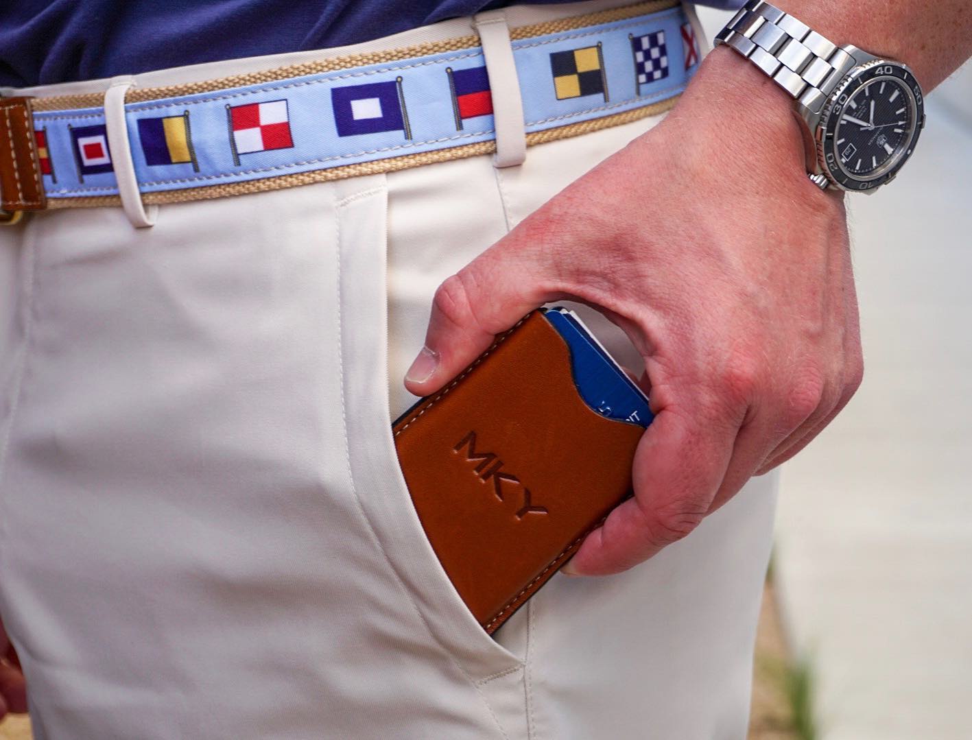 Close-up of a hand easily accessing a monogrammed British tan leather money clip holding cash from the front pocket of white trousers, demonstrating its secure hold for cards and cash.